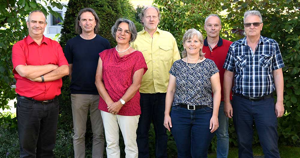 Sieben Personen (zwei Frauen, fünf Männer) stehen in der Natur vor Sträuchern